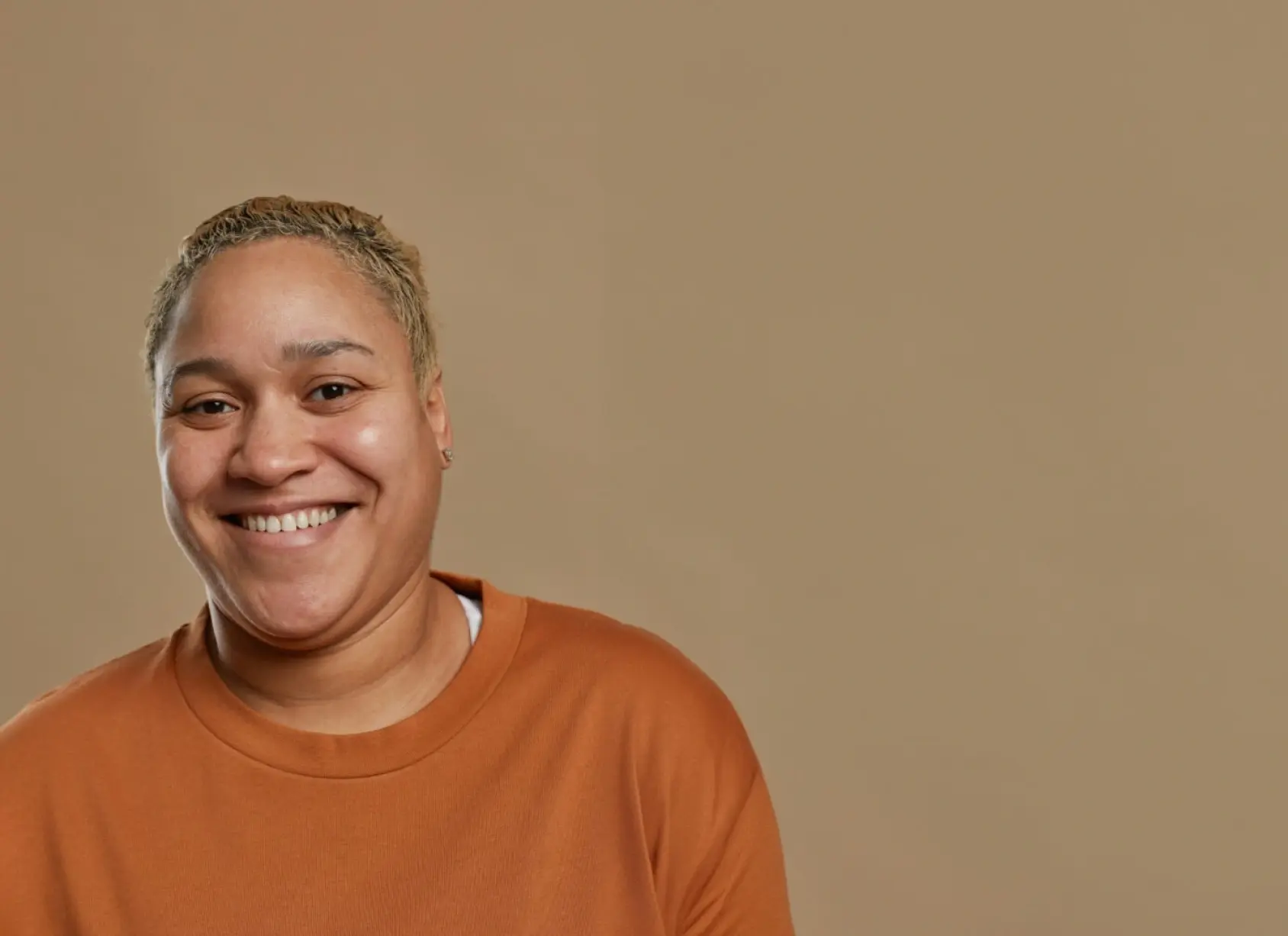 A person with short, light hair is smiling warmly while wearing an orange sweatshirt. The background is a solid, light brown color.