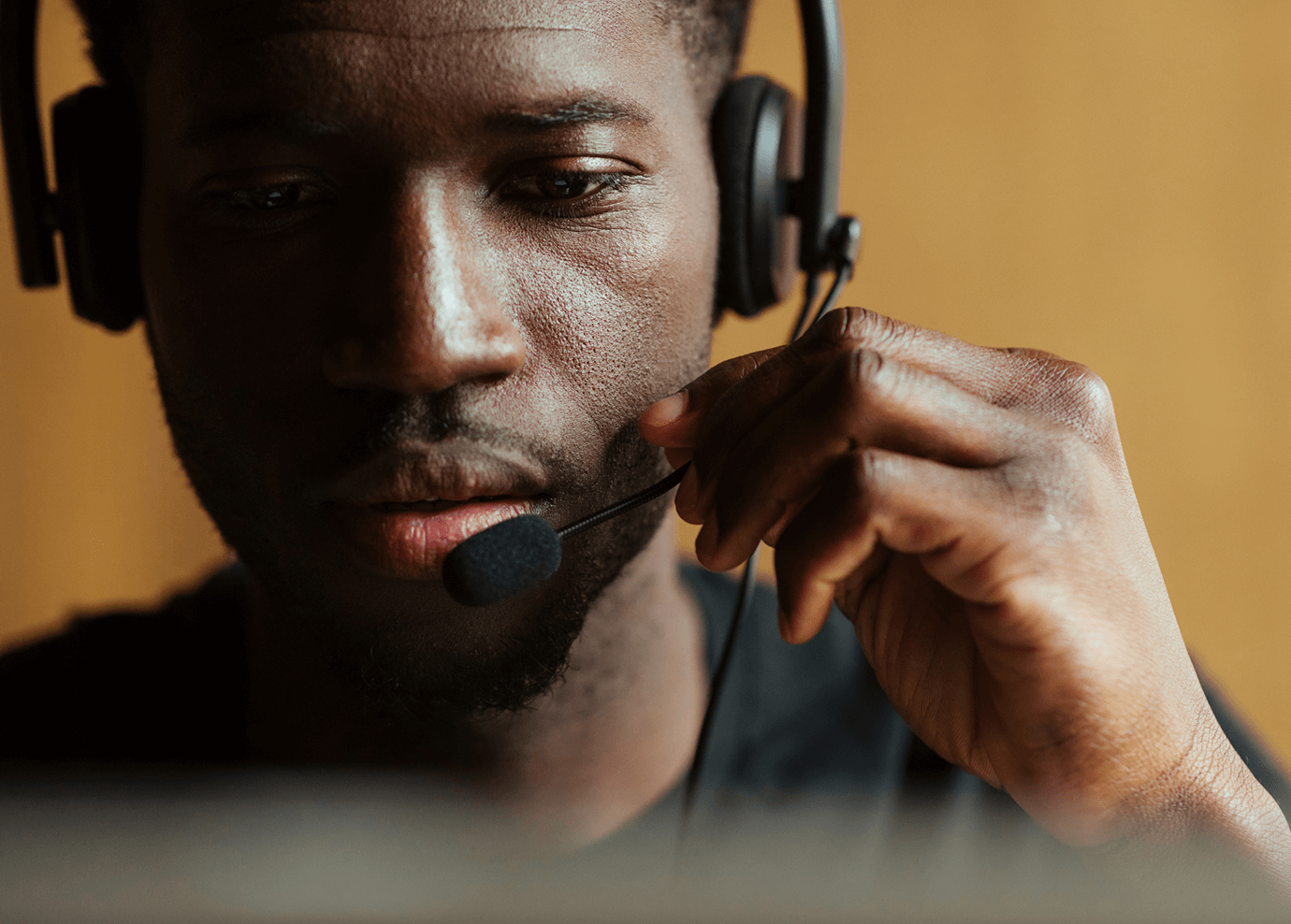 Close-up of a person wearing a headset with a microphone, appearing focused while speaking or listening, possibly working in customer service or a call center. The background is softly blurred.